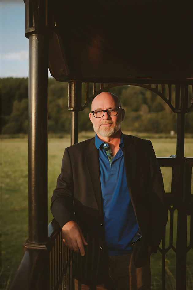 Jason Connery, stands in a suit near a large green field.